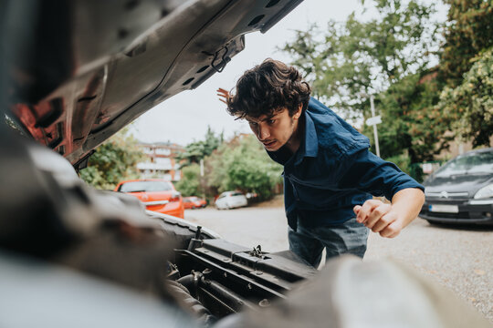 A young man intensely examines a car engine, showing focus and problem-solving skills in an outdoor location with parked cars and greenery.