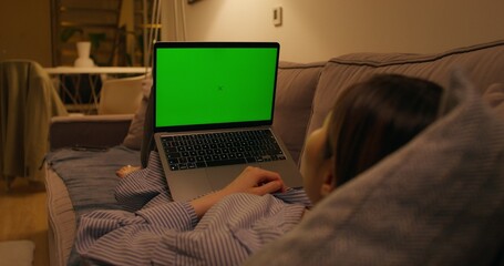 A woman uses a laptop with green screen lying on the couch in an apartment with dim lights