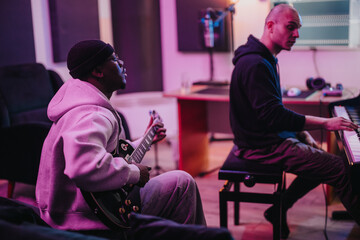 Two musicians create music in a vibrant recording studio. One plays electric guitar while the other plays piano, showcasing a creative collaboration in a modern setting.