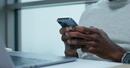 A man uses a mobile phone sitting at a table indoors. An open laptop is in front of him. Close-up of his hands, an unrecognizable person