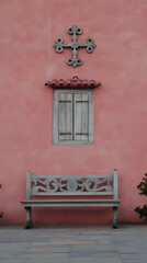 A weathered wooden bench sits below a shuttered window on a vibrant pink wall, adorned with a decorative cross.