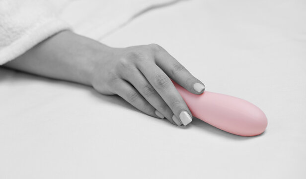 The girl is lying in a white bed. A woman holds a pink dildo in her hand. Toys for adults.