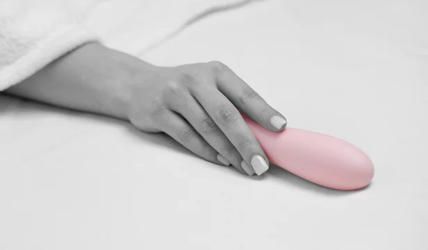 The girl is lying in a white bed. A woman holds a pink dildo in her hand. Toys for adults.