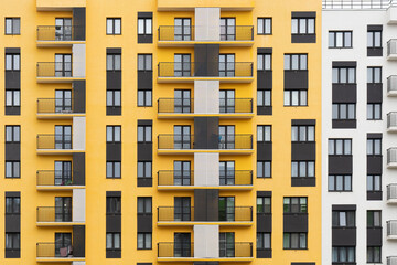 Modern apartment buildings with yellow and white facades, large windows, and balconies with metal railings, against a cloudy sky, front view