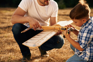 Messing around with broken toy. Father and son are with wooden plane on the field
