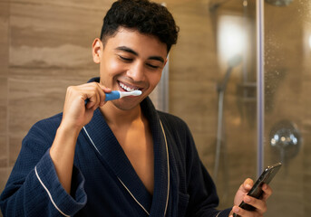 Young man brushing teeth in bathroom while checking phone in morning routine