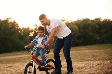 Obraz premium Teaching how to ride. Dad with son are with bicycle on the field outdoors