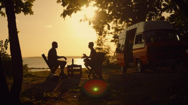 Romantic trip by motorhome in national park, silhouette of couple against sunset . Happy woman speaking funny stories to her boyfriend, calm family weekend in nature, enjoy solitude and peacefulness