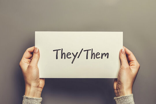 White sign with text 'they/them' held  by non-binary or genderfluid person