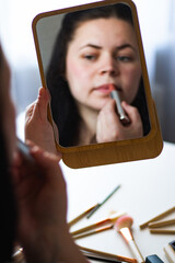 Vertical image of Millennial Caucasian woman doing self make up applying lipstick to lips while...