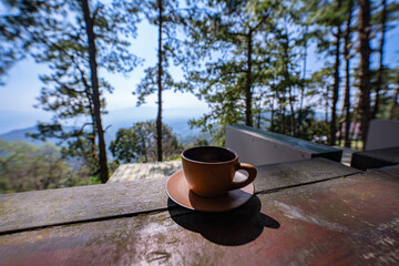 Hot coffee on a clear morning with pine trees lining the green forest under a blue sky. Green grass...