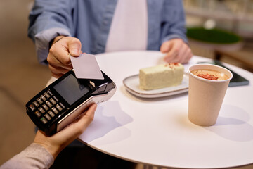 Close-up of hands making contactless payment in restaurant with credit card. Paper cup and dessert shown on table in background, creating cozy dining atmosphere