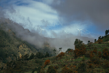 A picturesque view of mountains partially obscured by fog, featuring wind turbines standing tall against a dramatic sky, showcasing nature and technology in harmony.