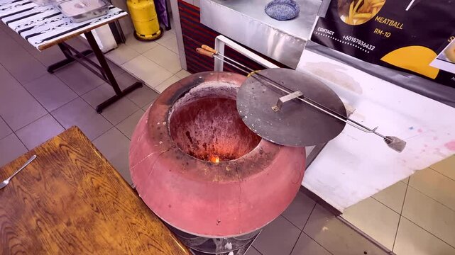 A traditional clay tandoor oven with an open lid, showing a fire inside, used for baking or grilling in a restaurant kitchen.
