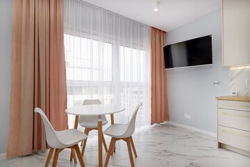 Salon w nowoczesnym apartamencie z beżowym  aneksem kuchennym z rudą jasno brązową pluszową sofą, biały stół z krzesłami pod dużym oknem