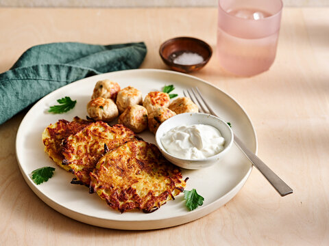 Golden brown potato latkes are plated with savory meatballs and a side of yogurt. Fresh cilantro and a glass of water enhance this appetizing meal setting
