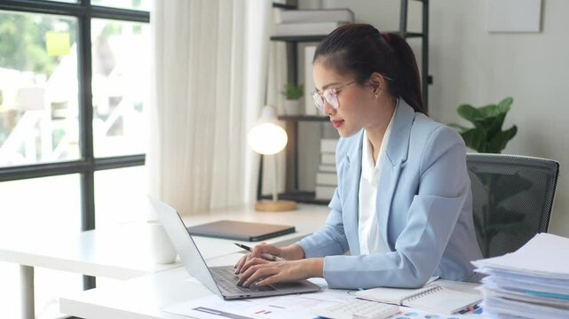A woman is sitting at a desk with a laptop and a stack of papers. She is wearing a blue suit and glasses