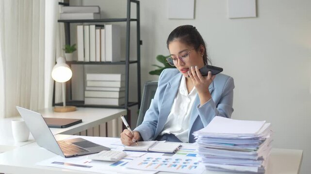 A woman in a business suit is talking on her cell phone while sitting at her desk. She is surrounded by stacks of papers and a laptop. Concept of productivity and focus