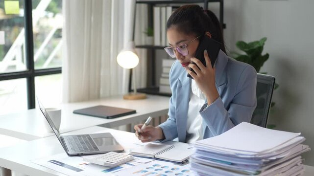 A woman is talking on her cell phone while sitting at a desk with a laptop and a stack of papers