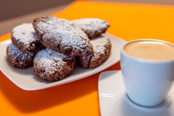 Pan dei Morti cookies stacked on a white plate, dusted with powdered sugar, paired with a creamy caffe macchiato on a vibrant orange background