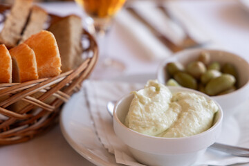 Traditional Mediterranean aioli served as an appetizer with fresh bread slices in a woven basket and a side of marinated green olives on a dining table setting