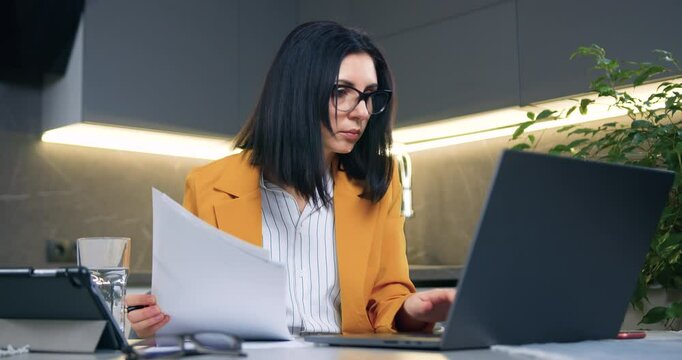 Business concept where beautiful concentrated considerate successful dark-haired businesswoman in formal wear sitting at home and working with computer and documents