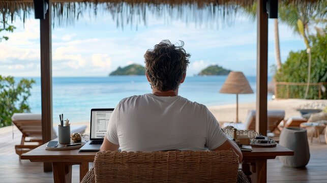 A person sits at a wooden table with a laptop, gazing at a serene beach view, surrounded by tropical decor and nature.