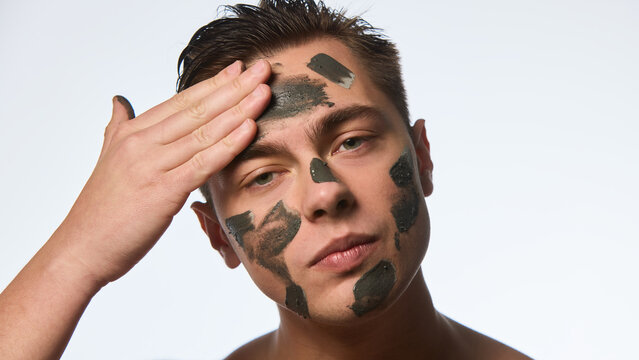 Close up photo of man with wet, brunette hair, doing facial procedures, applying black clay mask on skin against white background. Concept of selfcare and love, natural beauty, cosmetology.