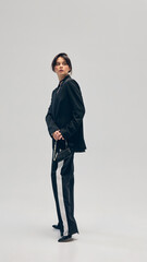 Full-length portrait of beautiful young woman in black suit, pants, blazer and small purse, posing...