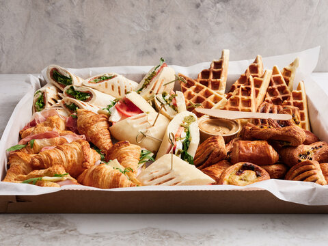 A colorful spread features a variety of savory sandwiches, flaky croissants, and sweet waffles presented in a catering box, perfect for a brunch event. The selection invites guests to indulge