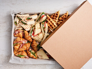 An assortment of sandwiches, fresh wraps, and golden waffles is arranged in a brown takeout box, ideal for sharing during a casual outdoor event or picnic