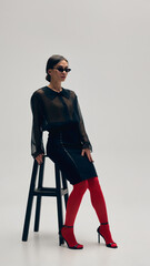 Graceful young woman in black blouse, skirt, red tights, heels and sunglasses posing on chair...