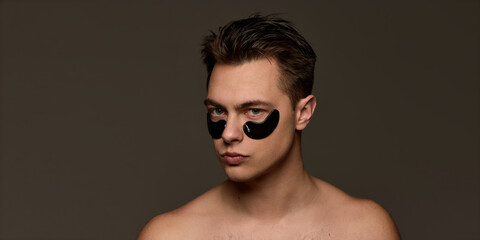 Shirtless young man with black eye patches, looks calm, embodying self-care and anti-fatigue skincare against brown studio background. Concept of selfcare and love, natural beauty, cosmetology.