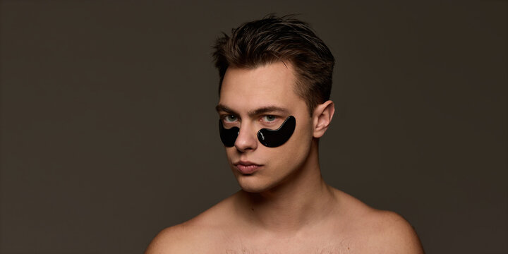 Shirtless young man with black eye patches, looks calm, embodying self-care and anti-fatigue skincare against brown studio background. Concept of selfcare and love, natural beauty, cosmetology.