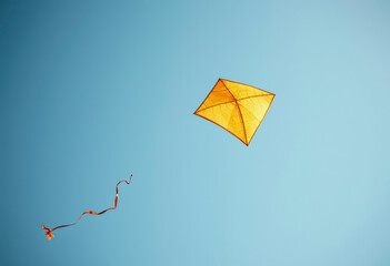 An orange kite with colorful ribbons dances in the wind against a bright blue sky, enjoyed by families at a park during daylight.