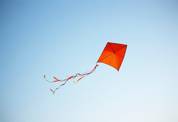 An orange kite with colorful ribbons dances in the wind against a bright blue sky, enjoyed by families at a park during daylight.