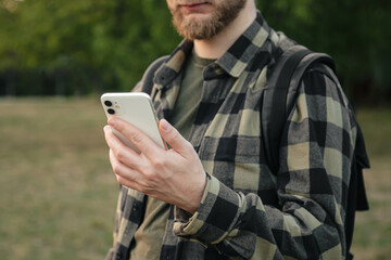 A man in a green shirt uses a white smartphone on a blurred background of nature outside, technology use concept, copy space.
