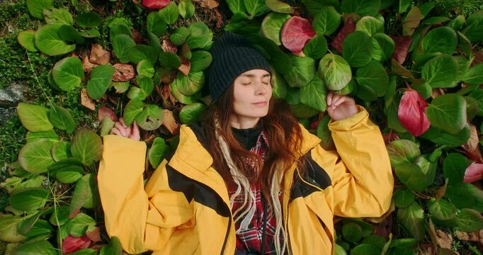 Young hiker wearing yellow jacket and beanie is lying on a patch of bergenia crassifolia with hands behind her head, enjoying the sun and the fresh air during a trekking day in the mountains