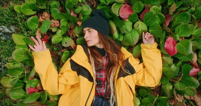 Young hiker wearing yellow jacket and beanie is lying on a patch of bergenia crassifolia with hands behind her head, enjoying the sun and the fresh air during a trekking day in the mountains
