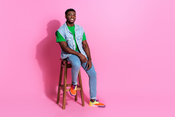 Young man in stylish denim vest and colorful sneakers sitting on a stool against a vibrant pink...