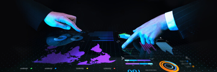 Business professionals collaborating over interactive digital table with global financial data, charts, and futuristic blue interface against dark background. Concept of business, data, engineering.