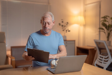 Focused mature man working remotely in a cozy home office, analyzing documents on a clipboard while using a laptop during the evening hours, embodying productivity and comfort