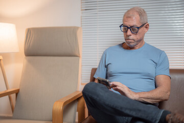 Relaxed senior man sitting comfortably on a cozy sofa in a well-lit living room, using his smartphone to browse the internet and enjoy leisure time in a peaceful atmosphere
