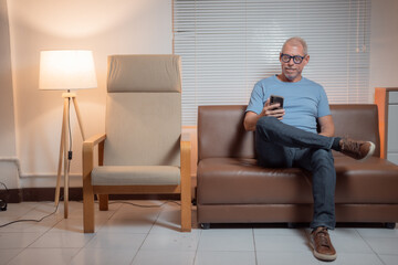 Fototapeta premium Happy retired man using smartphone apps, browsing the internet, reading news, chatting with friends, shopping online, and playing games while relaxing on a comfortable couch at home