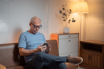Relaxed senior man using smartphone sitting on comfortable sofa at home, enjoying social media or messaging with family, browsing internet or watching videos