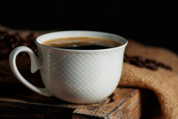 Close-up of hot coffee in a coffee cup With roasted coffee beans arranged on a table on a black and dark background.