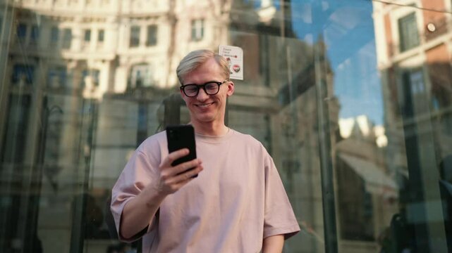 Smiling Caucasian man uses his smartphone to send money online via banking app with safety, standing near reflective glass building in vibrant city. ease, technology, and modern financial solutions

