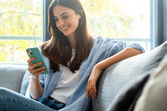 Young millennial woman sitting on a couch at home, casually dressed, smiling while using a smartphone to browse the internet, scroll social media, text friends, subscribe to online services - Powered by Adobe