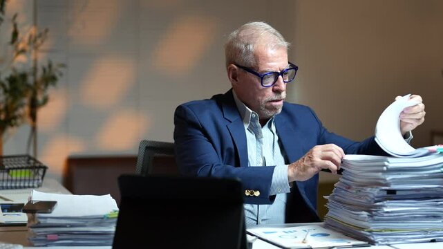 Professional businessman experiencing work-related stress. Overwhelmed by extensive paperwork while sitting at desk during late night overtime. Showing signs of workplace exhaustion and mental strain