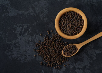 black pepper seeds in a wooden bowl on a black stone table, top view.
